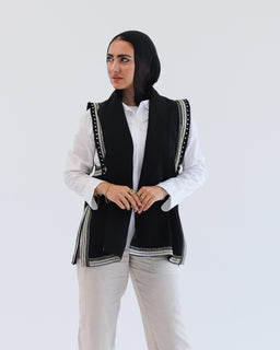 Product is a sleeveless vest with open front, styled over a white long-sleeve shirt. It has black body panels with decorative trim along the edges and arm openings, giving a tailored, layered look. The vest appears to be worn by a female model and suggests a unisex or women’s outerwear vibe with a modern, structured silhouette. Notable details include the black color, white/contrast stitching trim, and a slightly longer length that drapes over the hips. Material looks like a soft woven fabric, with a bold black-and-white contrast style.