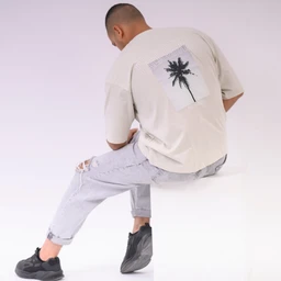 - Product: An oversized beige T-shirt with a palm tree graphic on the back.
- Audience: Unisex (model features a masculine fit and styling, but oversized and neutral tones suit both men and women).
- Notable details: Short sleeves, crew neckline, large back patch featuring a black palm tree image on a light rectangular background; paired with light-wash ripped jeans and dark sneakers in a casual, relaxed look. Material appears lightweight cotton or cotton-blend. Color palette is beige with a white-backed graphic.