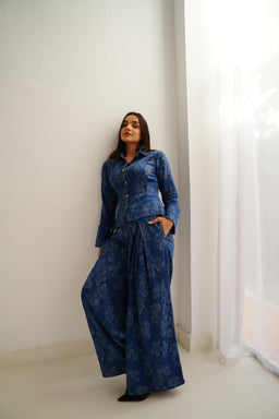 - A matching denim set featuring a long-sleeve button-front jacket and wide-leg pants with a blue floral pattern. The outfit is modeled by a woman standing in a bright, minimal white room, with one hand in a pocket and the other by her side.
- Intended audience: women's sizing and styling suggested by the cut and fit, with a chic, relaxed silhouette. Notable attributes: indigo blue washed denim, delicate floral print, tailored jacket with pockets, and wide-leg cropped-length pants.