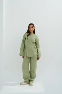 - What the product is: A two-piece linen loungewear set consisting of a wrap-style top and loose-fitting pants in olive green.
- Intended audience: Women (modeled by a female with a relaxed, oversized fit).
- Notable attributes: Soft olive linen fabric with a slightly textured surface, long sleeves with wide cuffs, a wrap-style top with a side tie, and wide, relaxed pants with a gathered or elasticized waist. The look is casual, comfortable, and easy-going, styled with light-colored sneakers.