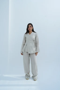 - Product: A matching linen set consisting of a wrap-style top with a V-neck and long, wide sleeves, paired with loose, straight-leg pants. The top features a side-tie/hip wrap detail giving a structured, asymmetrical silhouette; the pants have a relaxed fit with a soft, drapey fall.
- Intended audience: Women's.
- Notable attributes visible: light beige/stone color, linen-like fabric with a natural, matte finish, soft structure, and a minimalist, contemporary style. The model is wearing slip-on flat shoes, adding to a relaxed lounge-wear or resort look.