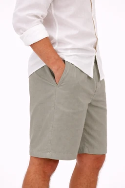This photo shows a pair of light olive linen shorts with a relaxed, straight fit and a flat front. The model appears to be male, wearing a white button-down shirt with rolled sleeves, suggesting a casual/summer men’s look. Notable attributes include the breathable linen fabric, pale olive color, mid-length short, and side pockets with a clean, minimal waistband.