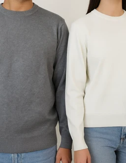 A round-knitted sweater in solid colors, shown on a male-presenting model (gray) and a female-presenting model (white), highlighting a unisex or versatile cut with classic crew neck. The gray sweater has a slightly heathered tone, while the white version appears smooth and clean, both featuring ribbed cuffs, collar, and hem. The styling is casual, paired with blue jeans, emphasizing everyday wear.