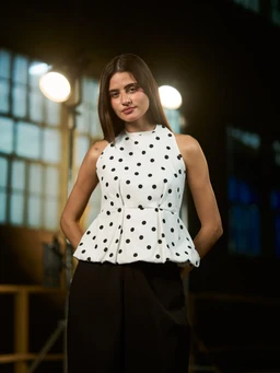 - A sleeveless white top with black polka dots, featuring a high neckline and a peplum waist that flares at the hips. The fabric appears structured, with a fitted bodice and a voluminous pleated peplum hem.
- Intended audience: women's.
- Notable attributes: white fabric with evenly spaced black polka dots, halter-like high neck, sleeveless, peplum silhouette, and a fitted waist creating a flared peplum bottom. No branding visible.
