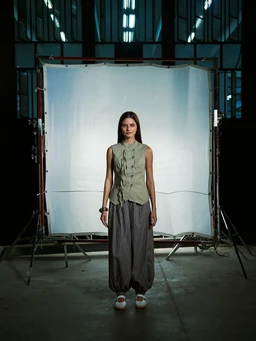 - Product: A sleeveless waistcoat or vest with a sculpted, tailored edge design in a light green/olive tone. 
- Audience: Appears unisex or potentially leaning toward women's fashion, judged from the model and styling (fitted vest over wide-leg pants, soft silhouette). 
- Materials/Style/Color: The vest has a structured, sculpted front with vertical lacing or seam details; fabric looks lightweight and matte. Paired with dark, loose-fitting, cropped trousers and white sneakers, giving a modern, utilitarian-meets-avant-garde style. The backdrop is a studio setup with a neutral light backdrop.