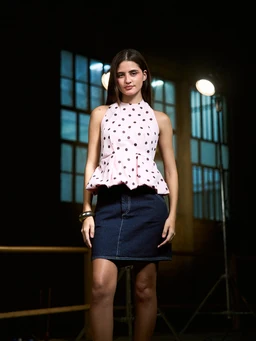 - What the product is: A women's sleeveless peplum top in light pink with dark brown polka dots and a high neck, paired with a dark denim mini skirt in the photo.
- Apparent audience: Women (based on the model, styling, and fit).
- Notable attributes: Light pink fabric with brown polka dots, halter/high-neck sleeveless silhouette, peplum waist flare, gathered/ruffled hem, paired with a dark blue denim mini skirt. Color palette is pink, brown, and denim blue; overall style is casual yet girly.