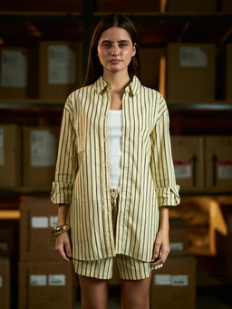 - What the product is: A yellow oversized striped poplin shirt worn over a white top, paired with matching striped shorts. The shirt is longline with a button-front and rolled cuffs.
- Intended audience: Appears to be unisex or women’s fashion, modeled by a female in a relaxed, oversized fit.
- Notable attributes: Light yellow tone with vertical brown and white stripes, lightweight poplin fabric, long sleeves with rolled cuffs, button-down collar, oversized silhouette, paired with matching striped shorts for a coordinated set.