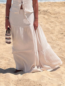 - Description: A flowing, light beige boho maxi skirt with a tiered, gathered silhouette. The fabric appears lightweight and crinkled, creating a soft, airy drape suitable for warm weather.
- Intended audience: Women's (based on styling, length, and fit on a female model).
- Notable attributes: Light beige color, boho tiered maxi style, ankle- or floor-length, airy textured fabric, paired with a matching top and accessorized with a woven bag. No branding visible.