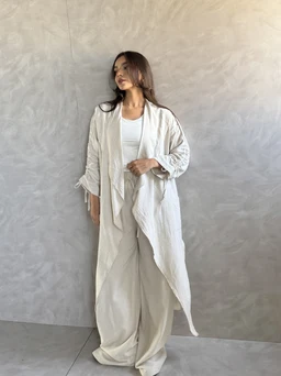 - What the product is: A long, cuffed cardigan in a soft beige tone, styled open over a white top with flowing, wide-leg pants. The cardigan features long sleeves with gathered cuffs and a draped, open-front design.
- Intended audience: Women (judging by the model, styling, and silhouette).
- Notable attributes: Light, creamy beige color; lightweight, textured fabric; long, relaxed silhouette with a cinched or ruched cuff detail; open-front cardigan with a draped, waterfall-style front.