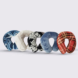 This shows a collection of U-shaped neck pillows arranged in a row, suggesting a unisex use for travel or lounging. They appear to be fabric-covered, filled with soft fiber, and feature a variety of printed patterns and colors including blue tones, cream, black and white floral, light blue, and red plaid. The notable attributes are the plush textile cover, rounded neck-support shape, and the fiber fill.