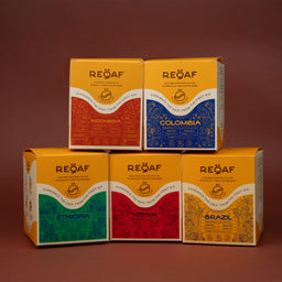 - A set of six small square coffee gift boxes, stacked in two rows (three on the bottom, three on top). Each box features a bright yellow top with a colored label area and bold country name (Indonesia, Colombia, Ethiopia, Kenya, Brazil), indicating a multi-origin coffee collection.
- Intended audience appears unisex, based on generic packaging and presentation, with vibrant, artisanal label styling. Notable attributes: bright yellow packaging, each box uses a different vivid color block and country-themed design, and the brand "RE{Q}AF" (no brand guessing beyond what is visible). Material and texture suggest cardboard gift boxes. Style is modern, colorful, and premium gift-box presentation.