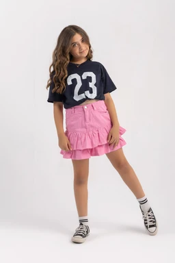 A layered pink denim skirt with tiers and ruffle details, paired with a navy oversized T-shirt featuring the number 23. The styling suggests a girls’ or young girls’ audience, based on the small model and playful color combination. Notable attributes include light-wade pink denim, tiered ruffle hem, and a casual, sporty-influenced look with black-and-white sneakers and striped socks.