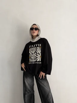 A black oversized T-shirt with a bold graphic on the front featuring white leaves and the words “FAITH OVER FEAR.” The outfit is styled for women, with a relaxed, loose fit and dropped shoulders, paired with dark washed wide-leg jeans and a light headscarf, plus sunglasses. Notable details include the oversized silhouette, monochrome palette, and graphic print, suggesting a casual streetwear look.