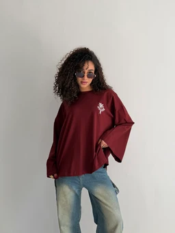 A loose, oversized maroon T-shirt with drop shoulders and wide sleeves worn by a female-presenting model. The shirt features a small white graphic/embroidery on the left chest. The model pairs it with light-wash, distressed jeans and round sunglasses, giving a casual, relaxed fit look. Material appears to be soft cotton or cotton-blend, with a matte-finish knit. Color: deep burgundy wine. Style: oversized short-sleeve T-shirt with wide, tumbled hem and relaxed silhouette. Audience: women's or unisex.