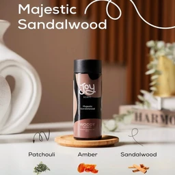- What the product is: A small, cylindrical bottle of Joy scent labeled “Majestic Sandalwood,” placed on a wooden coaster.
- Apparent audience: Unisex (neutral styling and placement; no gender-specific cues).
- Notable attributes visible: Dark amber bottle with a black cap and light pink/peach label; the scent name “Majestic Sandalwood” with component hints shown as Patchouli, Amber, Sandalwood at the bottom; soft, warm indoor background with decorative objects.