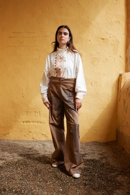 - Description: A woman wears a white, long-sleeve button-up shirt with subtle palm embroidery along the front placket and chest area, paired with high-waisted, wide-leg brown leather-look pants.
- Intended audience: Women's.
- Notable attributes: Cream/white shirt with light pink palm embroidery, relaxed, oversized sleeves with cuffs; high-waisted, loose-fitting brown trousers with a wide leg; neutral, earthy color palette. The look is vintage-inspired and artisanal in style.