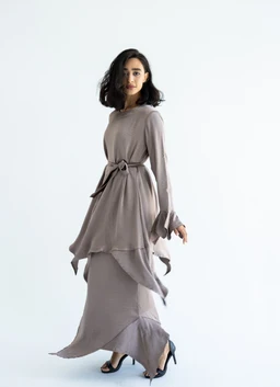A satin nightwear set featuring a soft taupe or dusty lilac color with a long-sleeve, high-neck top and a matching layered, asymmetrical skirt. The look is styled for women, with a waist-cinching belt tied in a bow to define the figure. Notable attributes include the silky satin fabric, flowy cascading hems, and a coordinated maxi-length silhouette.