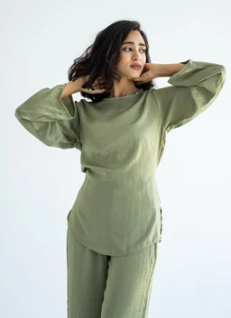 An olive green silk set featuring a matching long-sleeve top and relaxed trousers. The model appears to be a woman, styled in a loose, comfortable silhouette with voluminous sleeves and a soft, drapey fabric. Notable attributes include the satin-like silk texture, muted olive color, and a casual yet refined loungewear/edgy athleisure vibe.