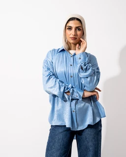 A light blue satin shirt with a relaxed, slightly oversized fit. It features long sleeves with gathered, ruched detailing and a button-front placket, styled here with a light hijab and pairing with dark wash jeans. The visible materials are glossy satin in a soft blue shade, with a casual, contemporary blousey silhouette suitable for women's fashion.