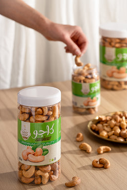 - This is a jar of roasted cashews (مكسرات كاجو) with a clear view of the nuts inside. The image shows a hand picking up cashews, plus another jar in the background and a small plate with loose nuts on a light wooden surface.
- Intended audience appears unisex (men and women) based on neutral packaging and presentation.
- Notable attributes: light brown cashews, transparent jar showing the nuts, white lid, green and white label design. The scene emphasizes a snack-style, premium quality look with a simple, natural color palette.