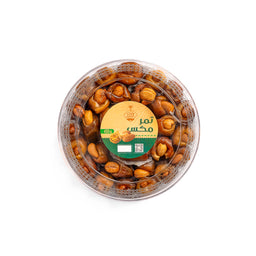 - A round, clear plastic盒 of mixed nuts glazed with a caramel-like syrup, arranged in a neat top-down view. The center features a round label in Arabic with green and orange accents, indicating a nut assort­ment product. The visible nuts appear roasted and glossy, with a mix of almonds and other kernels. Intended audience: unisex (general snack/relish). Notable attributes: warm brown roasted color, glossy coating, transparent container, Arabic labeling.