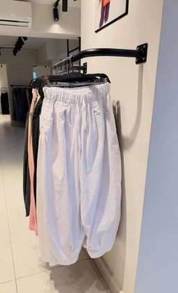 - Product: Balloon pants hanging on a rack inside a store fitting area.
- Audience: Women or unisex (based on the cut and styling seen; elastic waistband and loose, voluminous silhouette with gathered ankle resembles women's or unisex fashion).
- Notable attributes: Light pastel lavender/soft lilac color; oversized, drop-crotch balloon silhouette with elastic waistband and tapered, gathered cuffs; made from a lightweight, slightly shiny fabric.