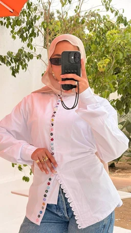 - Product: Daisy Shirt. The photo shows a white, long-sleeve button-front shirt with decorative multicolored flower-like buttons along the placket and a scalloped edge trim along the front opening and hem. It is styled with a light pink hijab and sunglasses, suggesting a modest, casual, warm-weather look.
- Audience: women's clothing (unisex style cues leaning feminine from the hijab styling and body fit).
- Notable attributes: white cotton or cotton-blend fabric, long sleeves, scalloped edging, colorful round buttons along the front, relaxed yet structured fit, paired with casual denim.