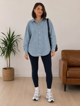 A light-wash denim shirt jacket with a relaxed, oversized fit and long sleeves. The model appears to be a woman, styled casually with dark leggings, chunky white sneakers, and a black backpack; cut is tunic-length with a button-front placket and a soft collar. Notable attributes: medium-blue denim, button-down placket, curved hem, and classic shirt-tail silhouette.