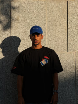- Product: A black short-sleeve T-shirt featuring a small graphic on the chest and a subtle logo on the left sleeve. The model is wearing a blue cap and a watch, standing outdoors against a light stone wall with strong side lighting creating shadows.
- Intended audience: Men’s (unisex styling appears suitable for adults).
- Visible attributes: Solid black fabric, round neckline, graphic patch on the left chest with a small colorful design, additional small text/logo on the left sleeve, casual oversized fit suggested by the model’s proportions. Cap is blue, and the color contrast highlights the shirt’s simple, everyday streetwear vibe.