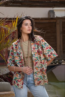 - Description: A short-sleeve, loose-fit shirt with a colorful tropical floral print worn over a beige top and light-wash jeans. The shirt appears linen or a linen-blend, featuring a relaxed, open-front style.
- Audience and styling cues: Women's sizing suggested by the model and the relaxed, casual styling with a pendant necklace and light denim.
- Notable attributes: Linen material, vibrant multicolor floral pattern on a light background, short-sleeve, oversized/boxy cut, worn over a neutral base layer.