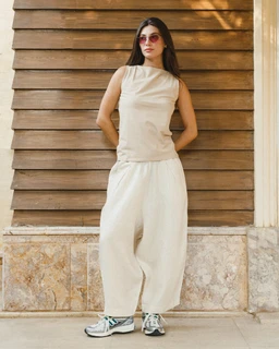 Product is a pair of linen balloon trousers styled for warm-weather wear. The visible model appears to be a woman (feminine fit) wearing a sleeveless, light beige top to match the trousers, with oversized, relaxed-cut pants gathered at the waist for a balloon shape. Notable visible attributes: natural linen-like fabric in a light cream/ivory color, wide, voluminous legs that taper slightly at the cuffs, elastic waistband, and a casual sneakers pairing. The overall look is airy, minimalist, and suitable for unstructured, everyday or resort/urban casual styling. Do not identify any brand.