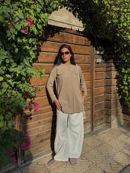 - The product is a long-sleeve, casual tunic/top in a soft beige-green tone, worn over wide-leg white pants. It appears designed for women, based on the styling, fit, and accessories (sunglasses, handbag).
- Notable features visible: lightweight knit or woven fabric, mandarin/flat collar, center seam lines, and relaxed, straight silhouette with side pockets. The overall look is minimalist and earthy, suitable for everyday wear in warm or mild weather.