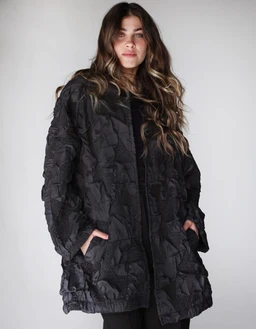 Product: Not so basic - Jacquard black jacket

- Apparent audience: women's (model styling, oversized relaxed fit, mid-length jacket)
- Notable features: black quilted jacquard texture, puffy padded construction, two side pockets, zip or hidden front closure, slightly oversized silhouette, long sleeves with a loose cuff. Material emphasis appears to be a padded, quilted fabric with a matte finish. Overall style is casual, outerwear for cool weather.