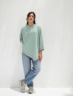 The product is a mint green asymmetrical shirt with long sleeves and a relaxed, draped fit. It appears aimed at women, judged from the styling and model. Notable details include a soft, lightweight fabric, a curved hem with orange-brown piping, and a loose, flowy silhouette paired with casual jeans and sneakers.
