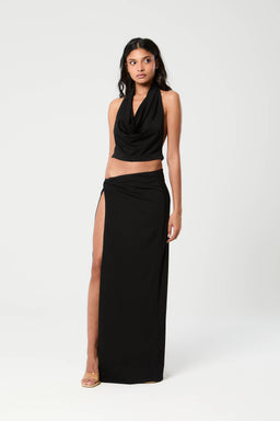 Description: A black two-piece set consisting of a draped halter-style crop top and a long, high-slit skirt. The model is a woman, styled with minimal jewelry and nude heeled sandals, conveying a chic, evening-ready look. Notable attributes visible: solid black fabric, halter neckline, draped front top, high waist skirt with a thigh-high slit, floor-length silhouette. Audience inferred: women's. Materials and fabric texture appear smooth and lightweight, with a soft drape. Color: black. Style: elegant, two-piece evening set.