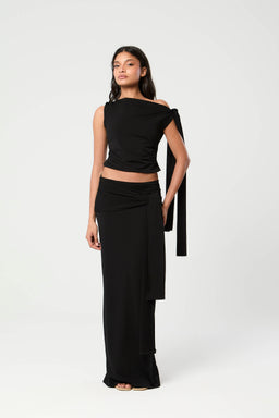 - A black outfit set featuring a cropped, off-shoulder top and a long, wide-leg skirt with a matching draped sash. The model is wearing a cape-like single-strap accessory hanging from the shoulder, creating a dramatic, layered look.
- Audience: women's fashion.
- Notable attributes: solid black color; cropped top with asymmetrical, off-shoulder neckline; long skirt with a wrap/draped sash detail; cape-like accent hanging from the shoulder; minimal sandals visible. Material impression suggests a smooth, flowing fabric with a slight drape.