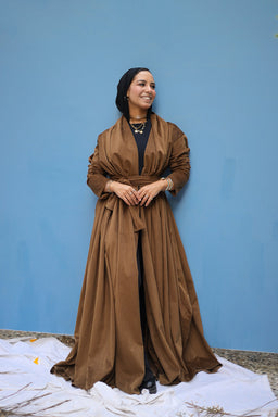 This image shows a long, flowing brown duster or abaya-style coat worn open over a black base outfit. The garment appears lightweight with a gathered waist and wide, dramatic sleeves, giving a voluminous silhouette. The model (adult woman) is styled with minimal accessories and a simple necklace, standing against a blue backdrop.