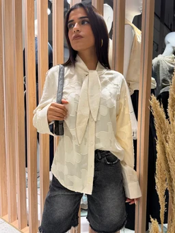 - What it is: A textured oversized shirt worn by a female-presenting model, styled with a necktie-like tie detail. 
- Audience indication: Women’s/ladies’ fashion, based on the feminine fit, necktie detail, and styling.
- Notable attributes: Off-white/cream color with a soft textured pattern; long sleeves; oversized relaxed fit; high-low or straight hem; paired with dark denim shorts for contrast. Material appearance suggests lightweight woven fabric.