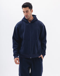 A navy blue zip-up hoodie with a full front zipper and a two-panel front pocket. The model appears male, wearing a relaxed, oversized fit with a drawstring-less hood and long sleeves. Notable materials look like a soft fleece or sweatshirt knit, presenting a solid dark blue color and casual sporty style.