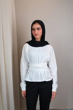- Product: Belted long sleeve crewneck top for women
- Audience: Women (based on styling and fit on female model)
- Notable attributes: White long-sleeve knit top with a gathered, belted waist creating a peplum-like silhouette; crew neckline; the model wears a black hijab, black pants, neutral backdrop. Material appears knit or jersey with a structured waist belt feature.