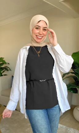 This is a women's layered top outfit featuring a black sleeveless tunic-style top worn under a white button-down shirt left open. The model appears to be a woman, accessorized with a beige hijab and a simple necklace, giving a casual, modest streetwear vibe. Notable materials and colors: black main top, white outer shirt, light beige hijab, denim blue jeans; the style is relaxed, with a cinched-in waist from a belt or tie.