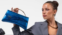 - What the product is: A blue metallic handbag with a curved, structured silhouette and a short top handle. The bag is shown being held up beside a model.
- Apparent audience and styling: Likely unisex to women; styled with a pinstripe blazer and chunky chain necklace for a modern, fashion-forward look.
- Notable attributes: Bright cobalt-blue metallic finish with a small logo emblem on the corner; rigid rectangular shape with a short handle and a subtle, glossy texture. No visible brand name beyond the logo.
