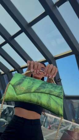 - What the product is: A small-to-medium green handbag with a metallic chain strap, held up by the model for display.
- Apparent audience: Women, judged from the cropped top and the handbag styling.
- Notable attributes: Bright green, tie-dye or marbled texture with darker green accents, gold-toned chain strap, compact satchel style with a flap closure and subtle stitched detailing. No visible branding on the bag itself.