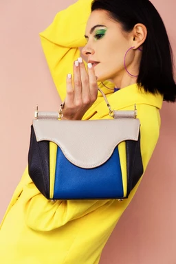 A small handbag with a structured, top-handle design. The model appears to be a female, suggesting the bag is aimed at women; styling is bold color-blocking with beige/cream flap, royal blue center panel, and dark navy side panels, accented by light hardware. Notable materials and colors include a smooth beige flap, blue and navy panels, and a bright royal blue and yellow color palette, with a compact, boxy silhouette and a curved flap detailing.