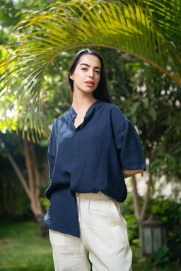 This is an oversized short-sleeve shirt in navy blue, modeled in an outdoor, tropical setting. The fit appears relaxed and slightly drapey on a female-presenting model, with a dropped shoulder line and a loose, boxy silhouette. Notable features include the textured, lightweight linen fabric, a subtle button-front placket, and mid-length sleeves; the color is a deep navy and the overall style is casual, breathable linen shirt.