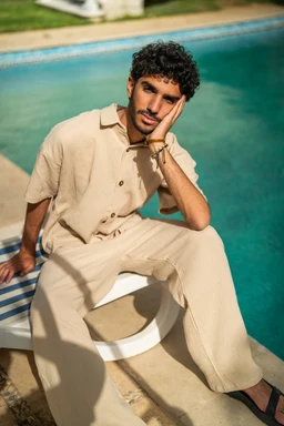 - A man sits poolside wearing an oversized beige shirt and matching wide-leg pants, giving a relaxed, casual look. The outfit appears to be a unisex or men's styling with a soft cotton texture, in a neutral beige color. Notable details include a short-sleeve shirt with a collar and front buttons, and the fabric’s light, breathable appearance suitable for warm weather.