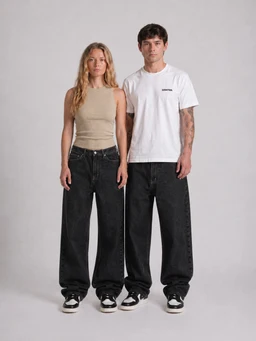- What the product is: A pair of relaxed wide-leg denim jeans in black, modeled by a man and a woman.
- Apparent audience: Unisex (fits both men's and women's styling, shown on a male and female model).
- Notable attributes visible: High-waist relaxed fit with very wide leg, black/dark wash denim, belt loops and front pockets visible, casual streetwear styling with sneakers. Clean, minimal top wear (tank top for the woman, white T-shirt for the man) to emphasize the jeans. Material appears to be sturdy denim.
