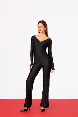 - A full-length black catsuit with long sleeves and a V-neck, fitted through the torso and hips, flaring slightly at the lower legs. The model is wearing black pointed-toe heels and a choker necklace, standing on a two-tone backdrop (white wall, red floor edge).
- Intended audience: women's, based on the styling, fit, and accessories.
- Notable attributes: sleek stretch fabric with a matte finish, deep V-neck, fitted silhouette, long sleeves, and flared trousers/le​gged cut at the bottom. Color: solid black. Style: form-fitting utility-meets-glam jumpsuit.