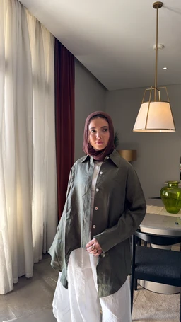 - What the product is: A round-neck, olive green button-front shirt-dress or long shirt, styled as a lightweight, oversized top.
- Apparent audience: Unisex or women’s, judging from the loose fit, styling, and model.
- Notable attributes: Olive green color, long sleeves, button-front closure, oversized silhouette, mid-thigh length with a curved hem, shirt collar. Visible environment includes a modern interior with a hanging lamp and neutral tones. Material appears lightweight cotton or similar fabric.