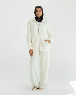 - What the product is: An ivory textured loungewear set consisting of a zip-front hoodie and matching wide-leg pants. The model wears a black headscarf underneath, with the zip partially open and hands in pockets.
- Apparent audience: Women's loungewear set, judging from the feminine fit, styling, and presentation.
- Notable attributes: Textured ivory fabric, full-zip hoodie with front pockets, relaxed wide-leg pants, soft and cozy casual style. Light, neutral tone with a comfortable, athleisure vibe.